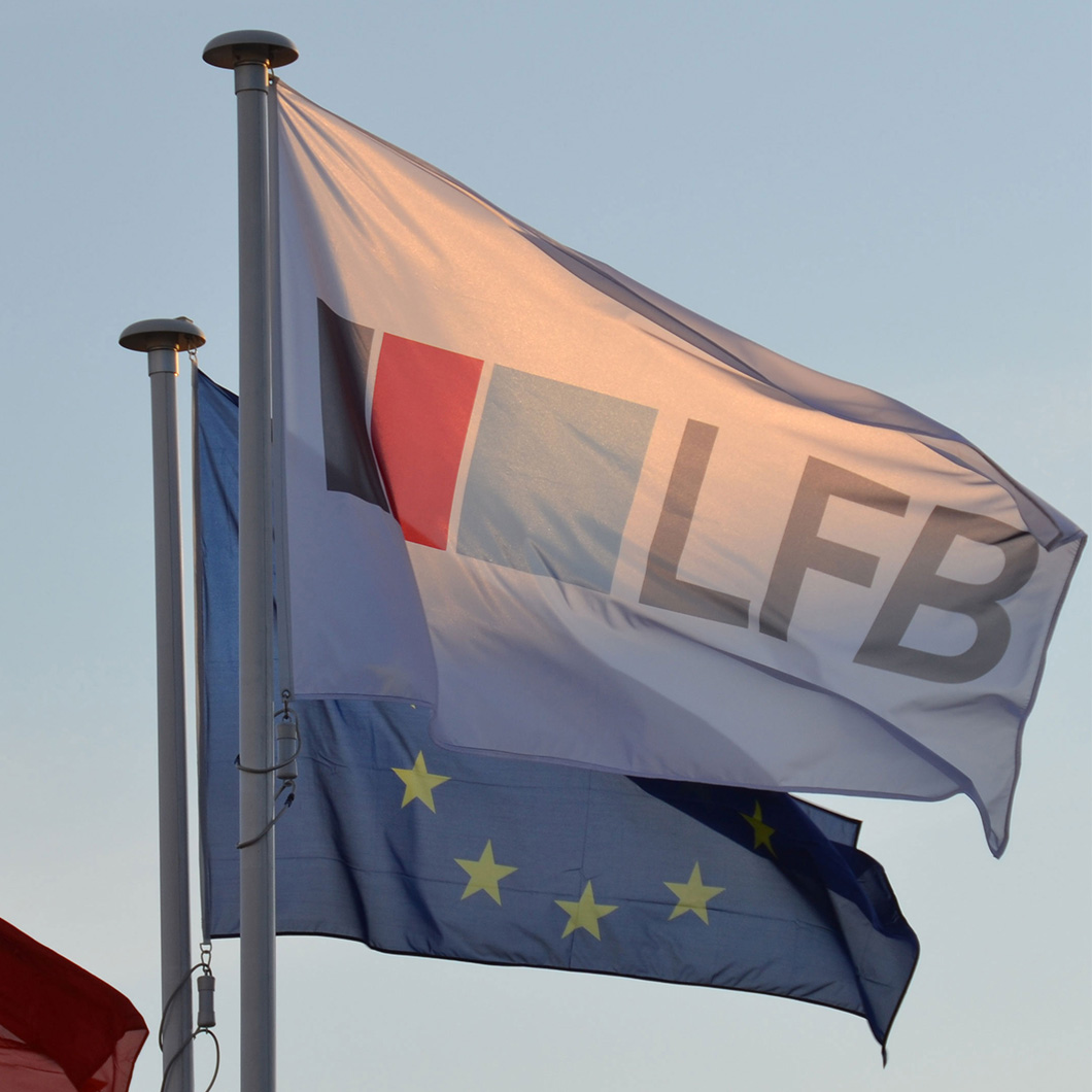Drapeau blanc du logo LFB flottant au vent sur un mât, avec un drapeau européen étoilé visible juste en dessous, sur fond de ciel bleu clair au crépuscule.