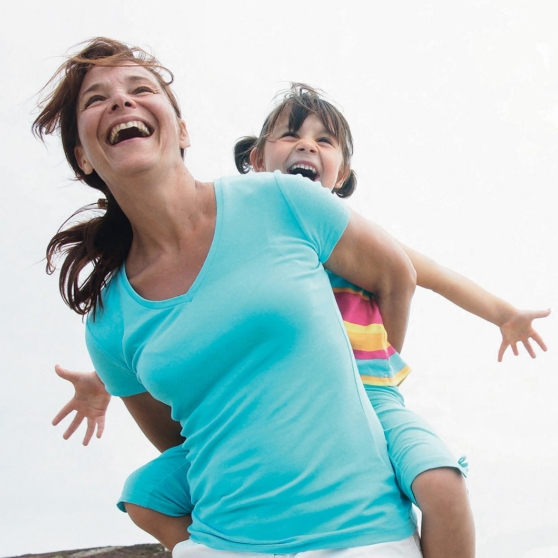 Photographie d'une femme souriante portant une petite fille sur son dos, toutes deux les bras écartés et riant, sur fond de ciel clair.