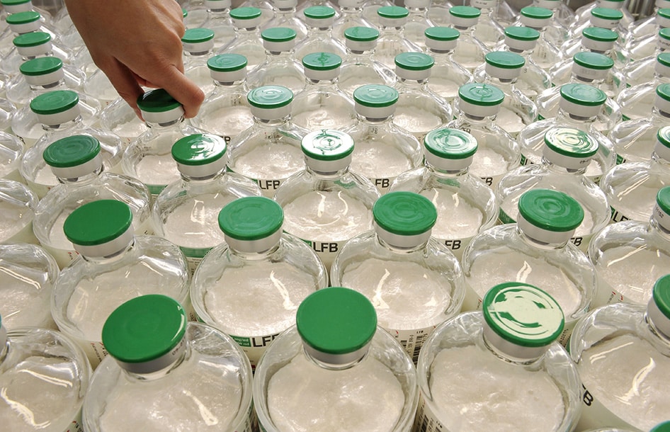 Photographie en plongée de rangées de flacons en verre remplis d'une poudre blanche, munis d'un bouchon vert et étiquetés LFB, avec une main s'apprêtant à saisir l'un des flacons.