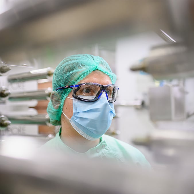 Portrait d'un opérateur en tenue stérile (charlotte, masque, lunettes de protection) dans un laboratoire de production pharmaceutique, entouré d'équipements en inox.