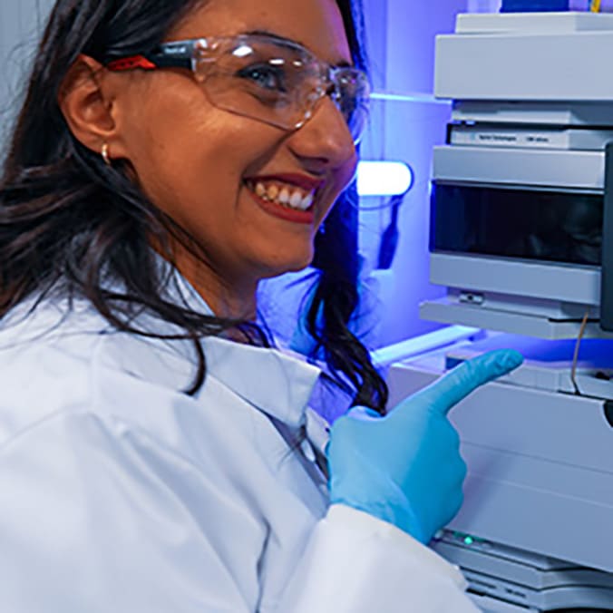 Photographie d'une femme souriante en blouse blanche et lunettes de protection, portant un gant bleu, désignant du doigt un appareil de laboratoire éclairé en bleu.