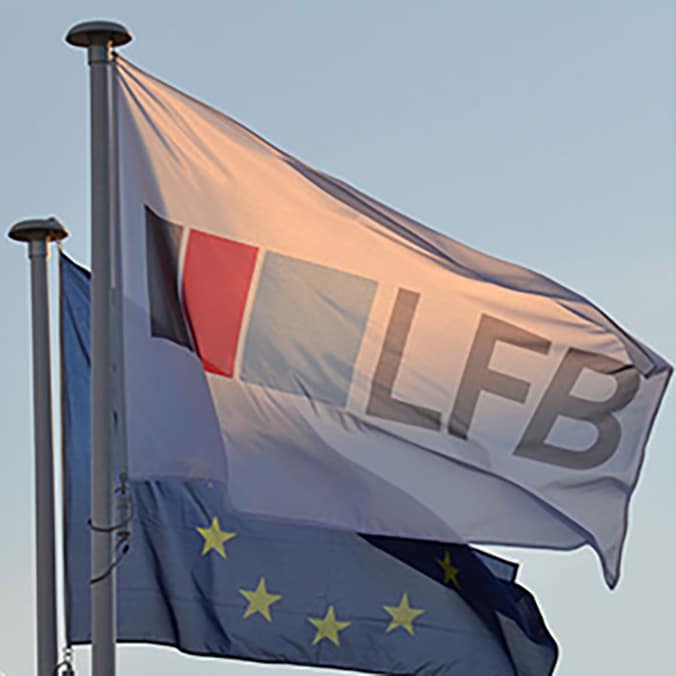 Drapeau blanc du logo LFB flottant au vent sur un mât, avec un drapeau européen étoilé visible juste en dessous, sur fond de ciel bleu clair au crépuscule.