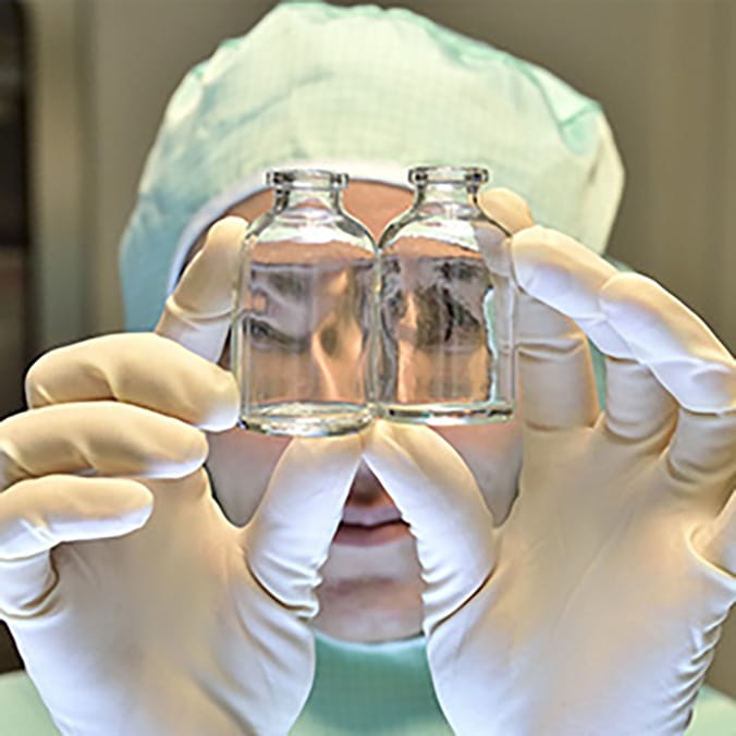 Gros plan sur une personne en tenue de laboratoire (charlotte, gants blancs) tenant deux flacons en verre transparents devant son visage, à hauteur des yeux.