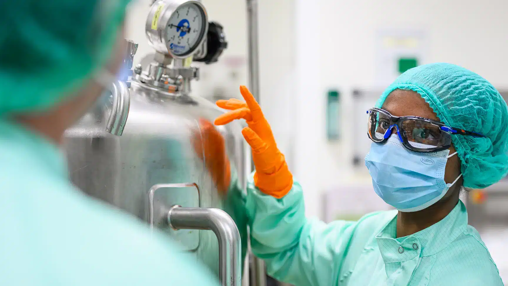 Deux opérateurs en tenue stérile (charlotte, masque, lunettes de protection, gants) travaillent sur une cuve de production en environnement pharmaceutique.
