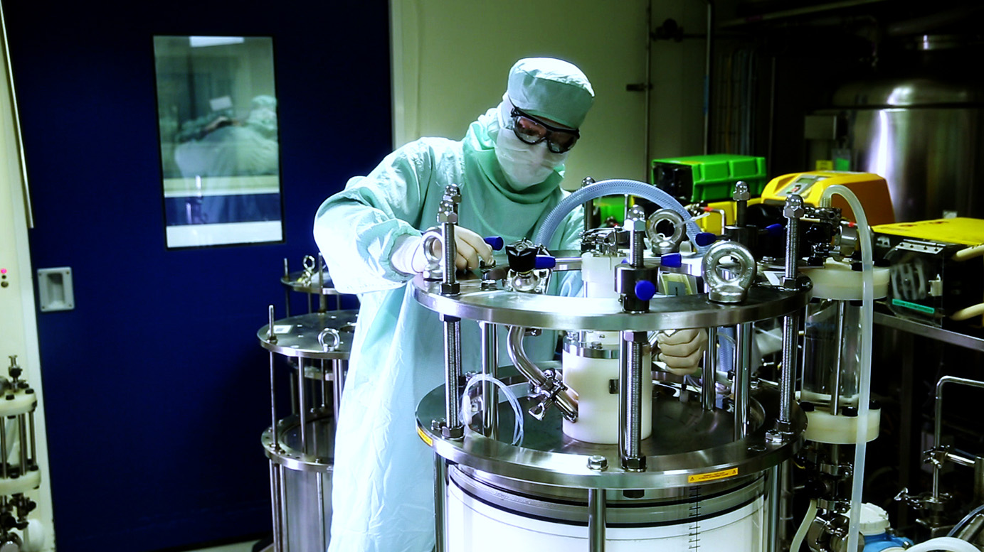 Un opérateur en tenue stérile complète (charlotte, masque, lunettes, blouse) manipule un équipement de purification en inox dans une salle de production pharmaceutique.