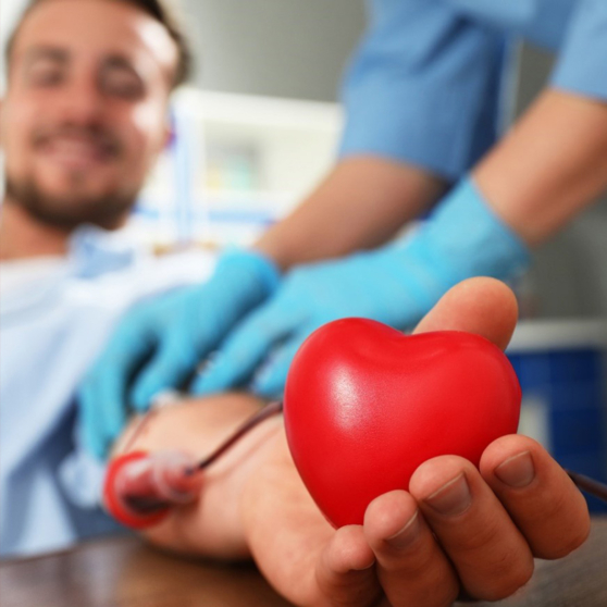 Photographie d'un homme souriant en cours de don du sang, tendant vers l'objectif un cœur en mousse rouge, tandis qu'une personne en tenue médicale s'occupe de son bras.