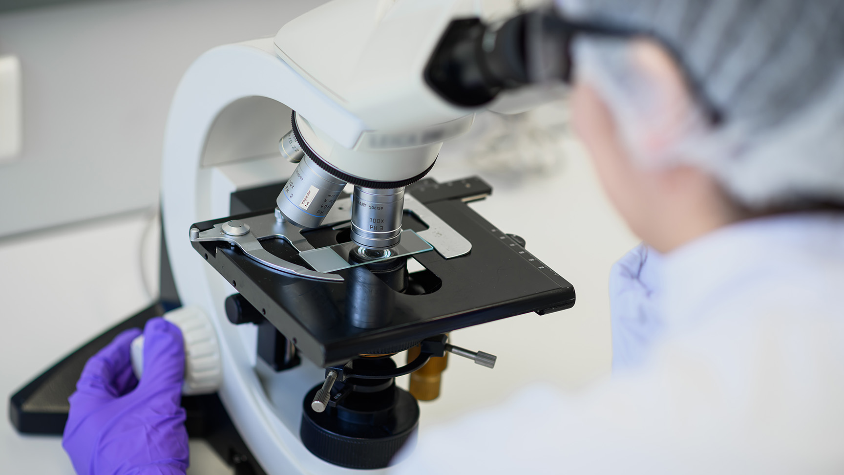 Photographie en gros plan d'un microscope de laboratoire, avec une personne en blouse blanche, charlotte et gants violets, penchée pour observer un échantillon.