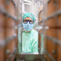 Photographie d'une personne en tenue de salle blanche (charlotte, masque, lunettes de protection, blouse verte), photographiée entre deux cuves industrielles en inox.