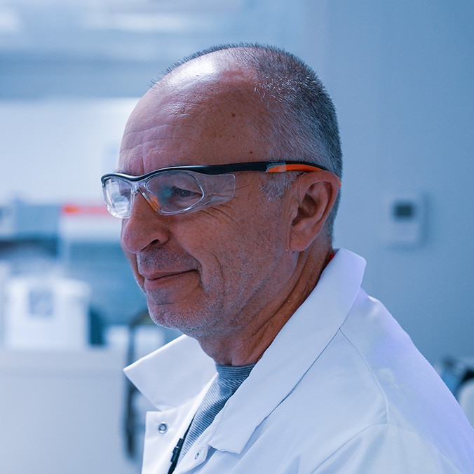 Portrait de profil d'un homme aux cheveux gris courts, portant des lunettes de protection et une blouse blanche de laboratoire, dans un environnement de laboratoire.