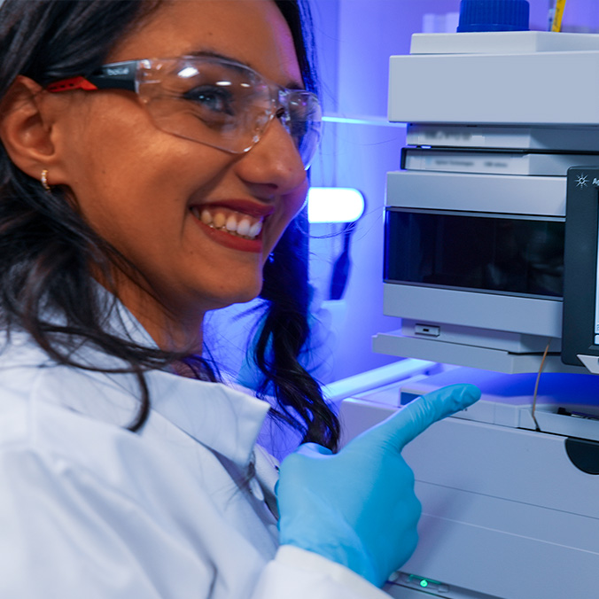 Photographie d'une femme souriante en blouse blanche et lunettes de protection, portant un gant bleu, désignant du doigt un appareil de laboratoire éclairé en bleu.