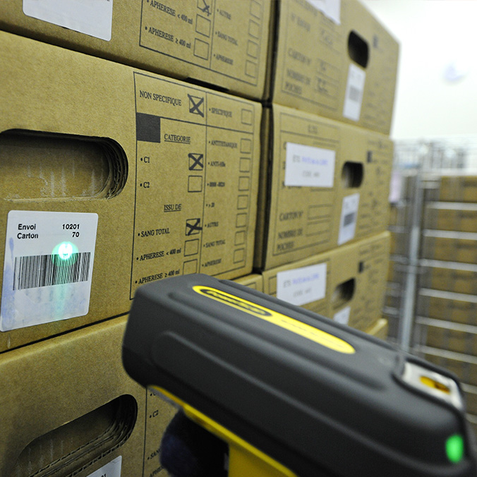 Un scanner code-barres lit l'étiquette d'un carton d'expédition parmi une pile de cartons de collecte de dons du sang, classés par catégorie et origine (sang total, aphérèse)