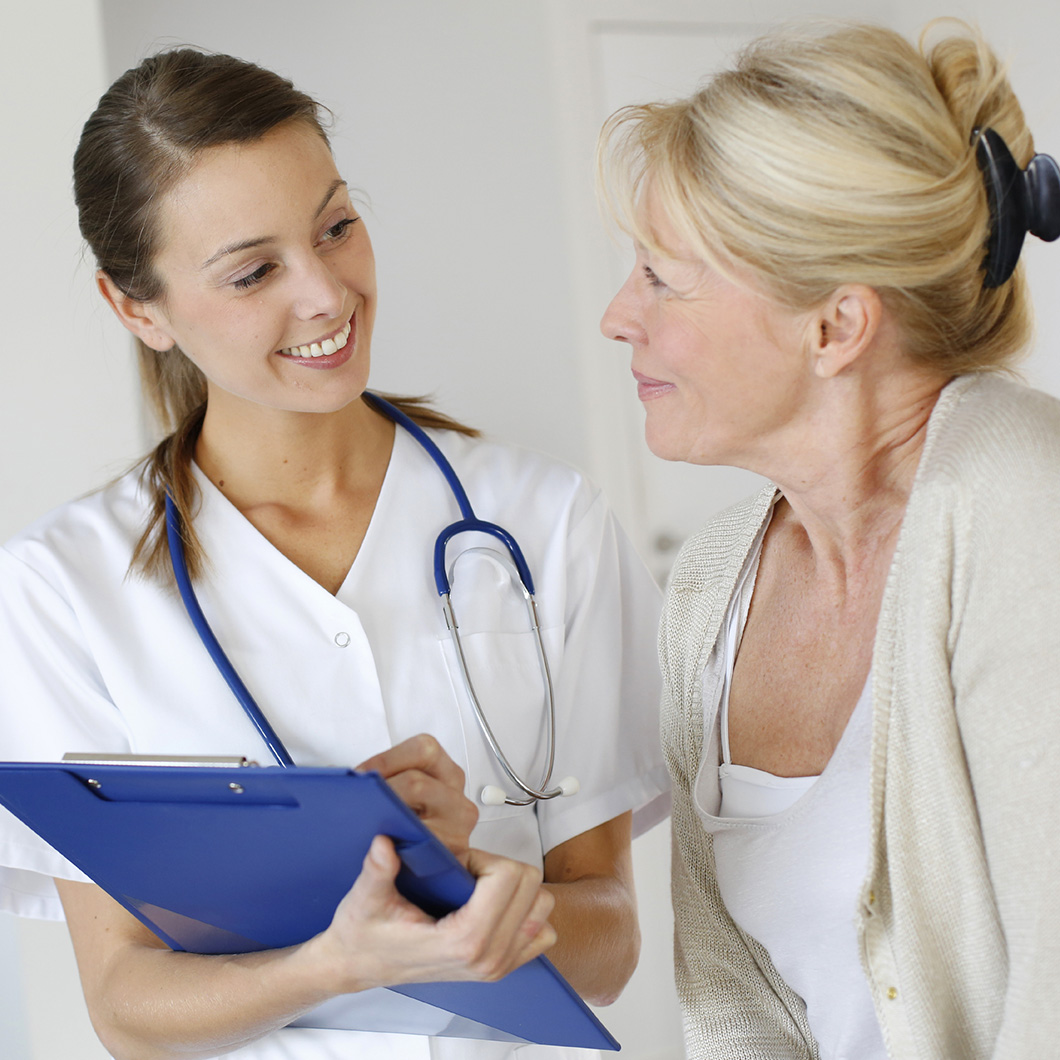 Photographie d'une infirmière souriante en blouse blanche, stéthoscope autour du cou, tenant un dossier, en échange avec une patiente aux cheveux blonds.