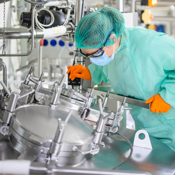 Un opérateur en tenue stérile (charlotte, masque, lunettes de protection, gants orange) manipule l'ouverture d'une cuve de production en inox dans un laboratoire pharmaceutique.