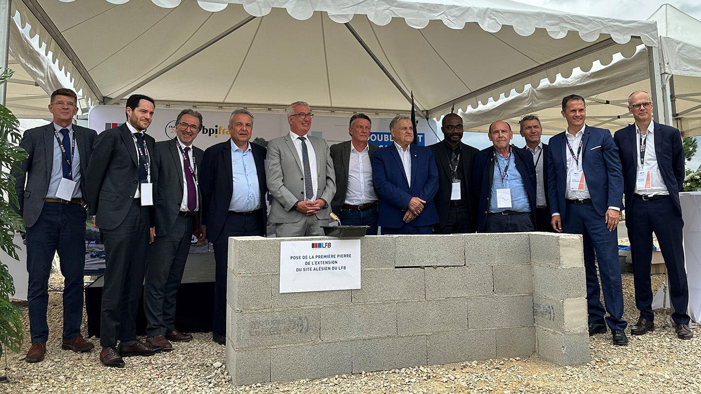 Groupe d'une douzaine de personnes en tenue professionnelle posant sous un chapiteau autour d'un muret de parpaings portant l'inscription « Pose de la première pierre de l'extension du site alésien du LFB ».