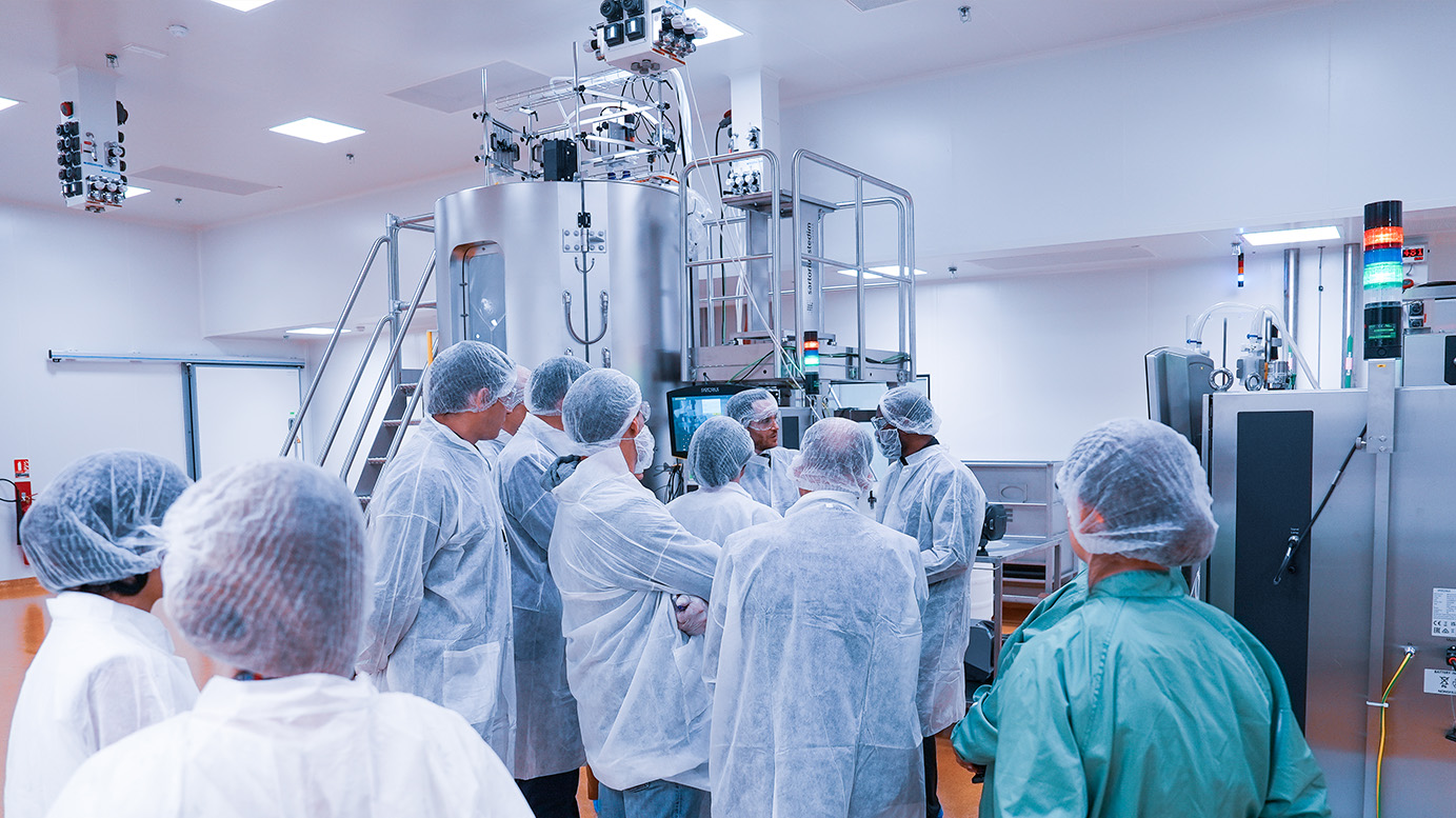 Groupe de personnes en tenue professionnelle réunies dans une salle de production pharmaceutique, vue de dos, devant une cuve inox équipée de câbles, capteurs et voyants lumineux.
