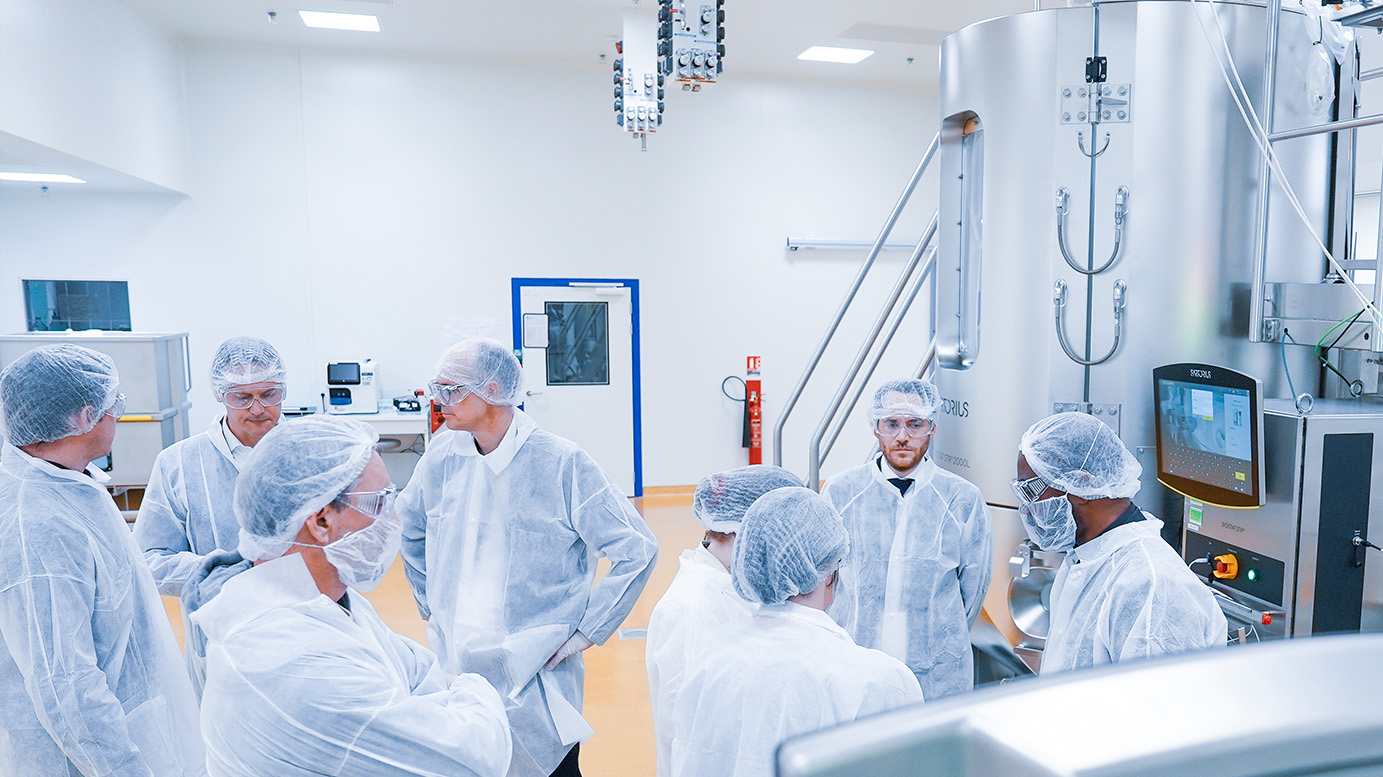 Groupe de huit personnes en combinaisons blanches, charlottes et lunettes de protection, réunies dans une salle de production pharmaceutique devant une cuve inox équipée d'un écran de contrôle.