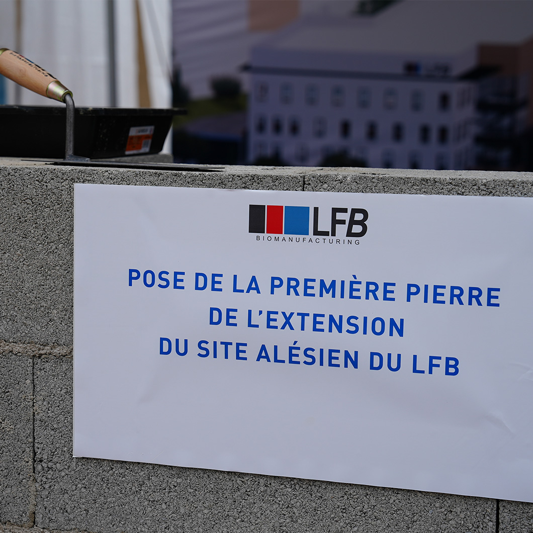Gros plan sur un muret de parpaings avec une pancarte : « LFB Biomanufacturing, pose de la première pierre de l'extension du site alésien du LFB ». Une taloche de maçon posée à côté, bâtiment en arrière-plan flou.