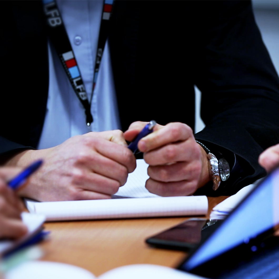Photographie en gros plan de deux personnes en réunion, tenant des stylos au-dessus de blocs-notes posés sur une table, avec un smartphone et un ordinateur portable à proximité. L'une des personnes porte un badge LFB autour du cou.