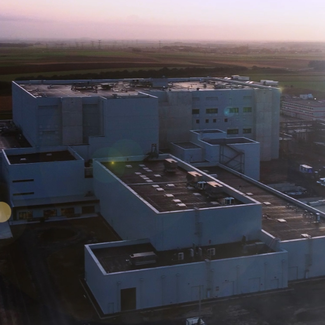 Vue aérienne de l'usine d'Arras. Un vaste complexe industriel aux bâtiments blancs, photographiée au crépuscule avec un ciel rosé, entourée de champs à perte de vue.