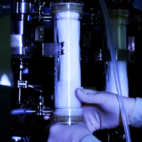Photographie en gros plan d'une main gantée manipulant une cartouche de filtration cylindrique translucide, composée de fibres fines, insérée dans un équipement de production, sous éclairage bleuté.