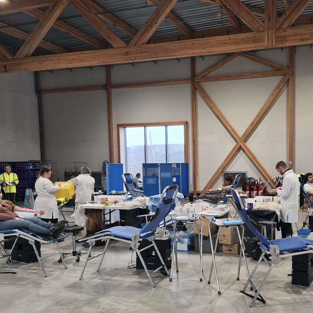 Intérieur d'un hangar à charpente en bois, aménagé pour une collecte de don du sang : plusieurs fauteuils bleus inclinables, donneurs allongés, personnel médical en blouse blanche s'affairant autour des tables de matériel.
