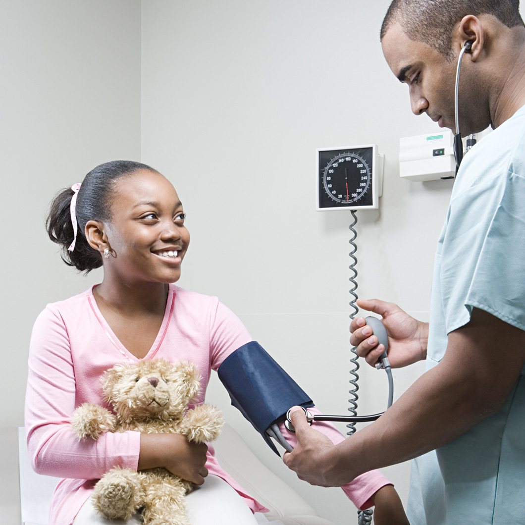 Photographie d'une fillette souriante tenant un ours en peluche se fait mesurer la tension artérielle par un soignant en tenue médicale.