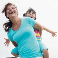 Femme portant une petite fille sur son dos, toutes deux souriantes et riant.