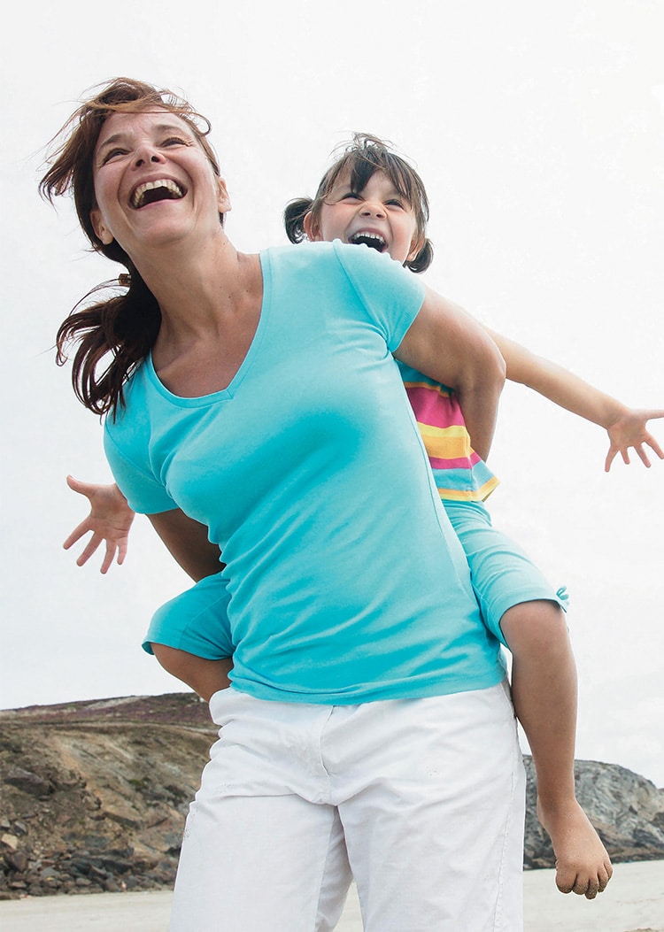 Femme portant une petite fille sur son dos, toutes deux souriantes et riant.